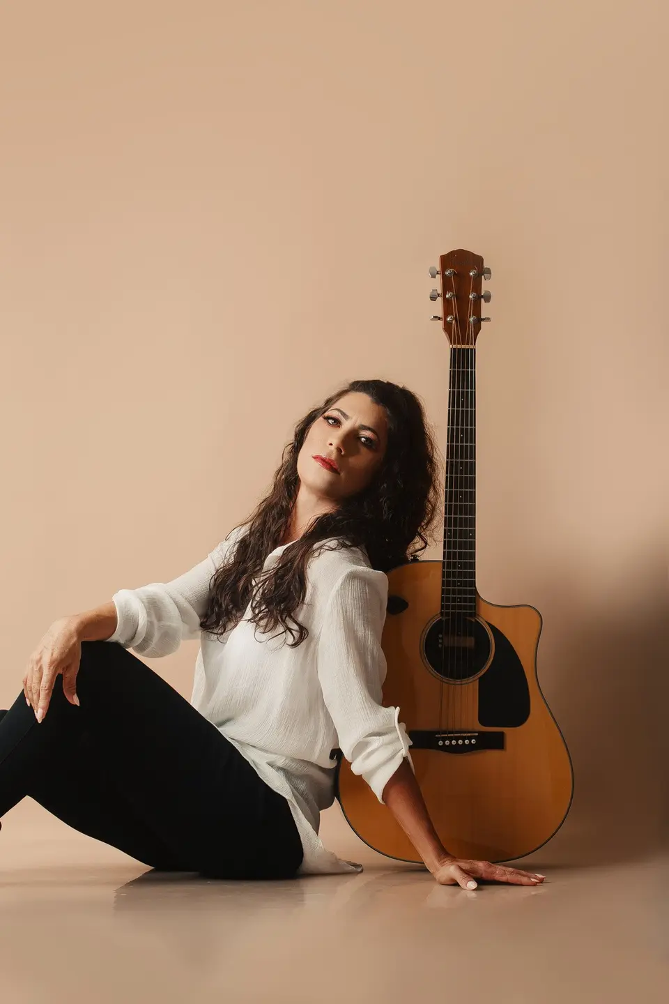 Lôids sentada no estúdio, de camisa branca, apoiada no violão
